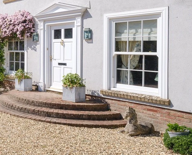 White front door with steps, flanked by two windows and potted plants, and a stone dog statue on gravel.
